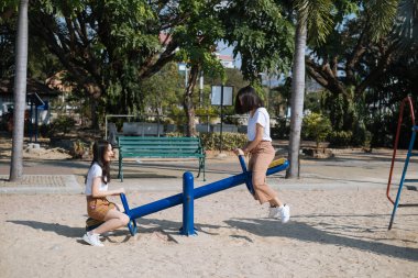 Beyaz tişörtlü iki kız gün ışığında parktaki tahterevallide oturuyor..