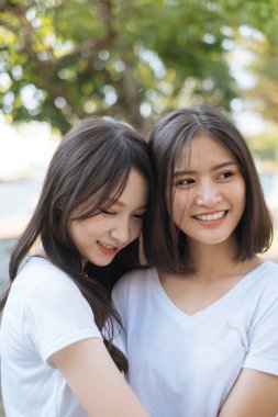 Parkta beyaz tişörtlü iki kız, uzun saçlı kız, kısa saçlı kıza sarılıyor..