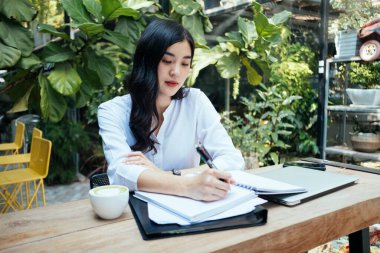 Genç Asyalı Taylandlı ofis kadını kağıt üzerinde çalışıyor, kalemle bir kitap yazıyor, açık havada bahçede..