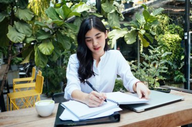 Genç Asyalı Taylandlı ofis kadını kağıt üzerinde çalışıyor, kalemle bir kitap yazıyor, açık havada bahçede..