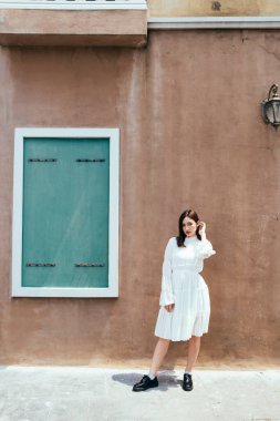 Beyaz elbiseli güzel, uzun saçlı kız yeşil pencereli ve lambalı kahverengi duvarın önünde duruyor..