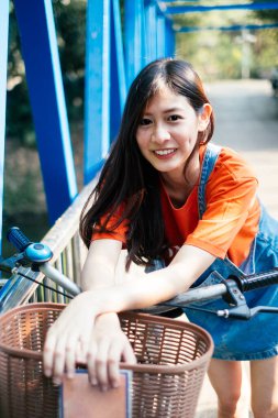 Uzun saçlı, turuncu tişörtlü ve kot pantolonlu kız mavi köprüde bisikletinin üzerine eğildi..