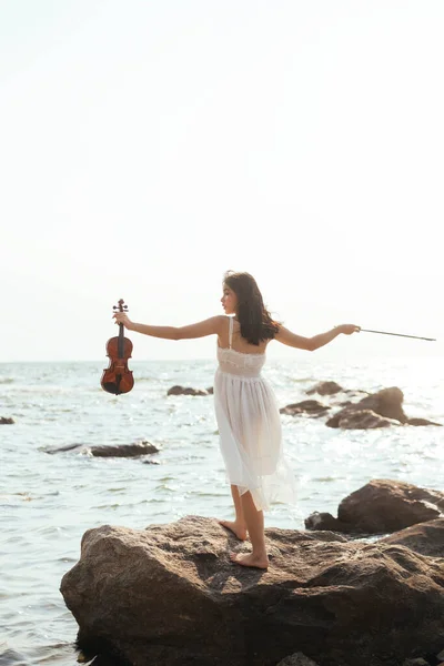 Black long hair woman in white dress standing on the rock with her violin and spread her arms out.