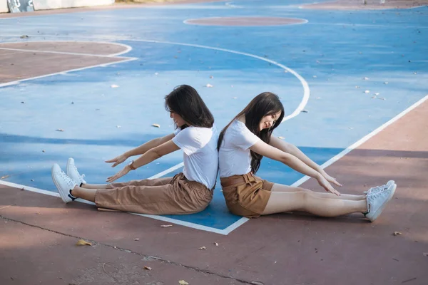 Two girls in white t-shirt sitting on basketball court with sttretched legs and try to touch their own feet.