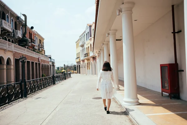 在古典风格的小镇上 一个身穿白衣的漂亮的长发姑娘走过大楼时的背影 — 图库照片
