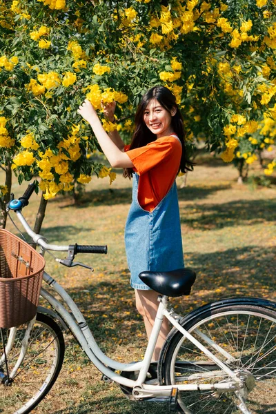 オレンジ色のTシャツとジーンズジャンパーの長い髪の女の子は自転車で庭で黄色の花を選ぶ — ストック写真