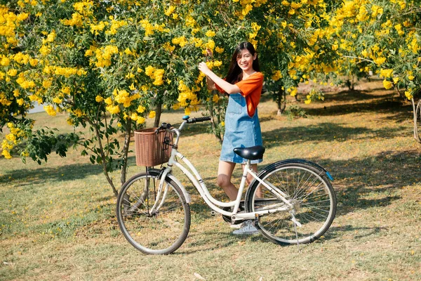 オレンジのTシャツとジーンズジャンパーの長い髪の女の子は 自転車 水平と庭で黄色の花を選ぶ — ストック写真