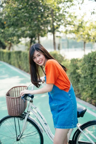 自転車レーンのツリーの横に彼女の自転車で立っている間 オレンジのTシャツとジーンズジャンパー笑いの長い髪の女の子 — ストック写真