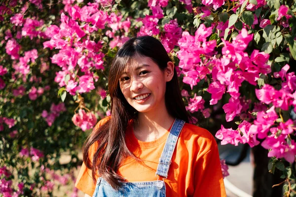 穿着橙色T恤和牛仔裤的长发女孩站在阳光下的粉色花冠旁边笑着的画像 — 图库照片