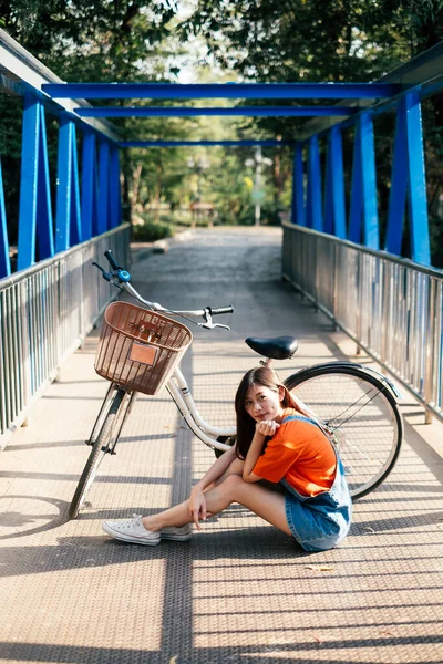 Long hair girl in orange t-shirt and jeans jumper sitting on the blue bridge with her bicycle, vertical.