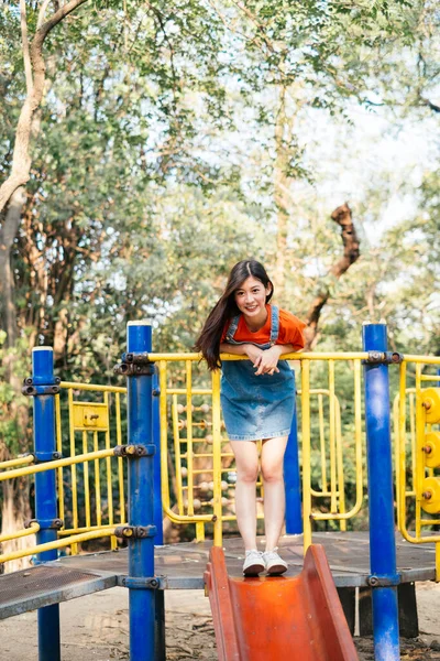 Long hair girl in orange t-shirt and jeans jumper standing on the top of playground slider, vertical.
