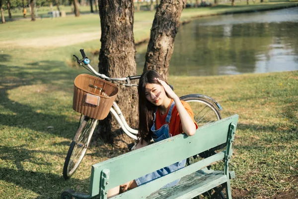 緑の公園の緑のベンチに座っているオレンジのTシャツとジーンズジャンパーの長い髪の女の子 日光に対する彼女の手のガードを上げる — ストック写真