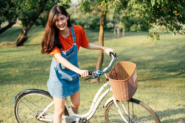 公園のフィールドで自転車と一緒に立っているオレンジのTシャツとジーンズジャンパーの長い髪の女の子 — ストック写真