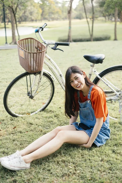 彼女の自転車で公園の芝生のフィールドに座っているオレンジのTシャツとジーンズジャンパーの長い髪の女の子 — ストック写真