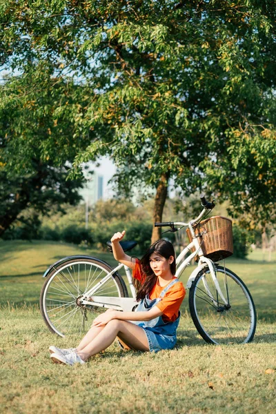 彼女の自転車で公園の芝生のフィールドに座っているオレンジのTシャツとジーンズジャンパーの長い髪の女の子 日光に対する彼女の手のガードを上げます — ストック写真