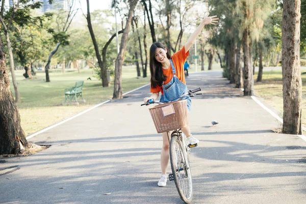 幸せな長い髪の女の子でオレンジTシャツとジーンズジャンパーは 公園内の道路に自転車に乗るのを停止し 空に彼女の手を上げる — ストック写真