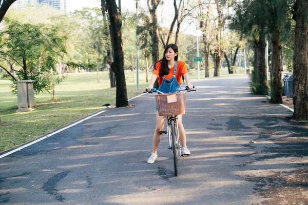 公園の道路上で自転車に乗るのを止めるオレンジのTシャツとジーンズジャンパーの長い髪の女の子 — ストック写真