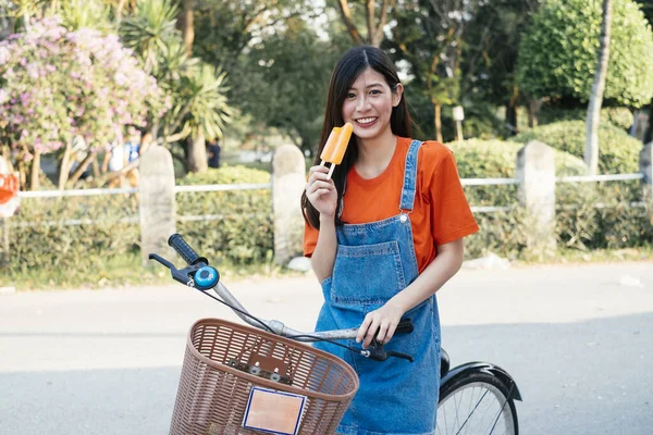 Long hair girl in orange t-shirt and jeans jumper show the orange ice cream on the bike, horizontal.