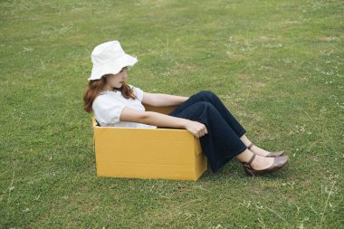 Genç Asyalı Taylandlı kadın hippi kıvırcık saçlı beyaz gömlekli yeşil park çayır tarlasında karton kutunun üzerinde oturuyor..