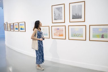 Mavi elbiseli, beyaz duvarda yürüyen ve sanat eserlerine bakan Asyalı güzel bir hippi kadın. Sanat galerisi.