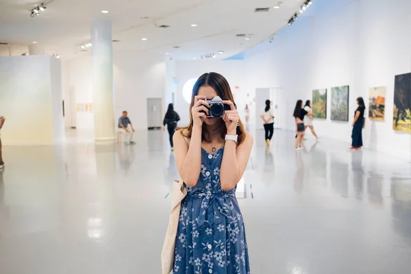 青のドレスを着た若いアジア系タイ人女性がアートギャラリーでカメラで写真を撮る — ストック写真