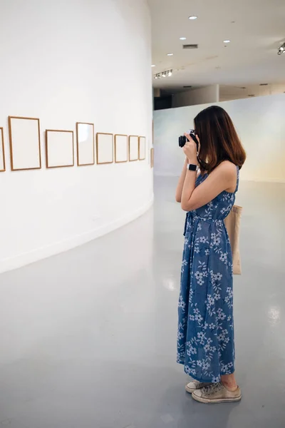 Young asian thai woman in blue dress taking picture with camera in artwork gallery.