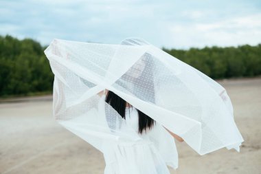 Açık havada beyaz peçeli Asyalı, uzun siyah saçlı bir kadın gelinin portresi. Rüzgarlı bir gün. Ülke tarafı arkaplanı.