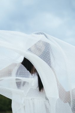 Açık havada beyaz peçeli Asyalı, uzun siyah saçlı bir kadın gelinin portresi. Rüzgarlı bir gün. Ülke tarafı arkaplanı.