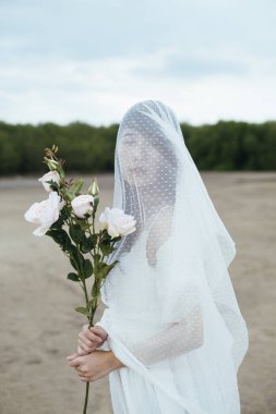 Beyaz gül çiçekli, açık havada duvağı olan Asyalı uzun saçlı bir gelinin portresi..