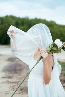 Beyaz gül çiçekli, açık havada duvağı olan Asyalı uzun saçlı bir gelinin portresi..