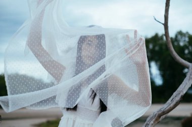 Genç Asyalı uzun saçlı kadın gelinin portresi vücudu beyaz peçeyle kaplıyor kuru ağacın önünde duruyor..