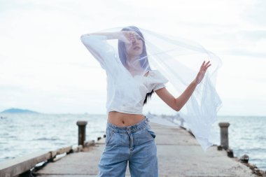 Deniz köprüsünde beyaz peçeli Asyalı uzun saçlı genç bir kadının portresi. Gömlek ve kot pantolon..