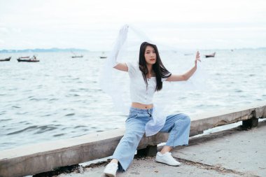 Uzun, siyah saçlı, beyaz peçeli genç Asyalı kadının portresi deniz köprüsünde oturuyor. Gömlek ve kot pantolon..