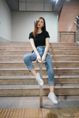 Tam boy moda portresi - Siyah gömlekli ve kot pantolonlu güzel Asyalı genç kadın yeraltı merdivenlerinde oturuyor.