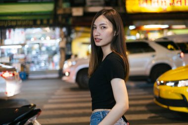 Moda portresi - Siyah gömlekli, kot pantolonlu, Asyalı güzel bir bayan geceleyin Bangkok 'ta karşıdan karşıya geçiyor..