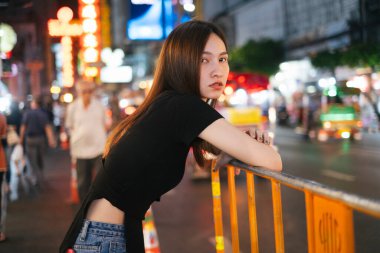 Moda portresi, siyah gömlekli ve pantolonu kaldırıma dayanmış güzel Asyalı genç bayan. Şehir gece hayatı.