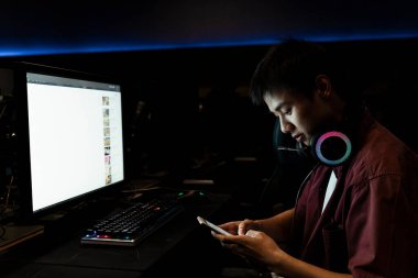 Bilgisayarın önünde oturan Asyalı oyuncu cep telefonu kullanıyor. İnternette sohbet ve sörf yapmak.