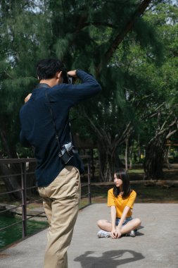 Erkek fotoğrafçı, parktaki küçük köprüde ayakta ve otururken genç bir kadın modelin fotoğrafını çekiyor..