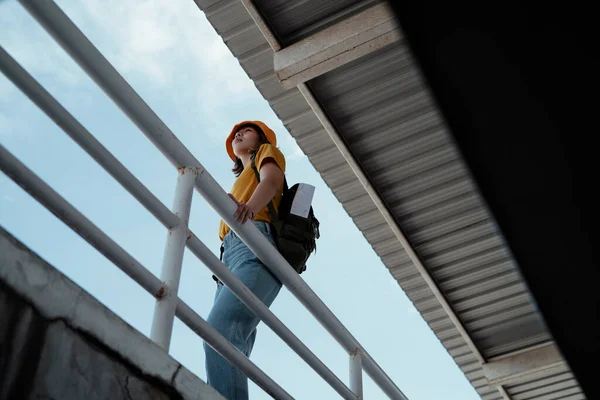 Low angle shot of yellow backpacker woman with green backpack standing at the overpass with roof and sky as a background.
