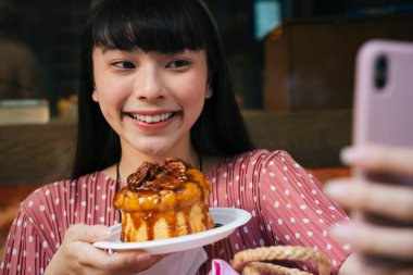Güzel Asyalı uzun siyah saçlı kadın kafe fırınında ekmek kekiyle selfie çekiyor..
