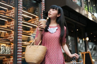 Güzel Asyalı uzun siyah saçlı, pembe elbiseli kadın Bangkok 'taki fırının önünde buz gibi soğuk kahvenin tadını çıkarıyor..