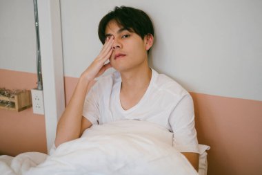 Beyaz tişörtlü, siyah saçlı bir adamın orta boy portresi. Yatakta otururken sağ gözünü kapatmış..