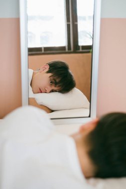 Siyah saçlı adam karın üstü yatar, yastığa sarılır ve aynadaki yansımasına bakar..