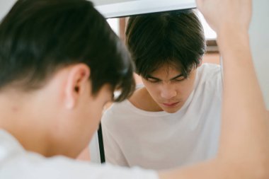 Siyah saçlı adam aynanın çerçevesini tutuyor ve yansımasının önündeki yere bakıyor..