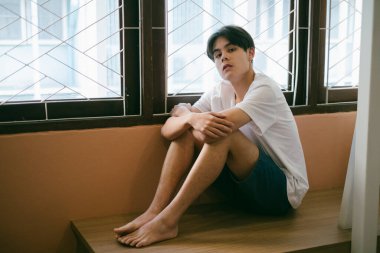 Kısa saçlı adam pencerede tek başına oturur, dizlerine sarılır ve kameraya bakar..