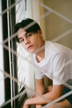 Beyaz tişörtlü adam pencerenin yanında oturuyor, boş kafayla dışarıya bakıyor..