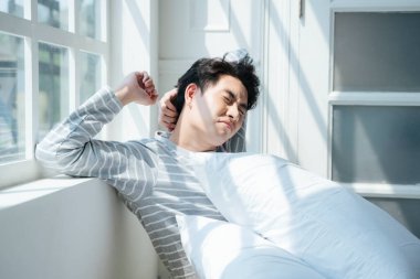 Sabah ışığıyla pencerenin yanındaki yatakta oturan bir adam acıyı hafifletmek için vücudunu esnetiyor..