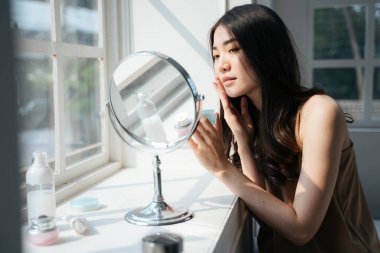 Dalgalı uzun saçlı kız yatak odasının penceresinin yanındaki aynanın önünde otururken yüzüne kremi sür..