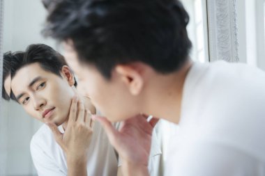 Beyaz tişörtlü bir adam aynada yüzüne bakar ve pezevenkleri bulur..