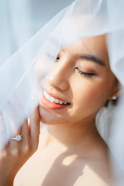 Pony tail blond hair woman finish dress up in the morning of her wedding day, standing nex to the window, cover herself with white veil and touch the window to show her wedding ring.